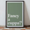 A framed wall art print featuring minimalist typography with the text "Fancy a cuppa?" in bold white letters on a muted green background, placed against a textured floor.
