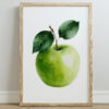 A framed watercolor illustration of a green apple with two green leaves, set against a white background, resting on a light-colored wooden floor.