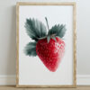 A framed watercolor illustration of a ripe strawberry with green leaves, set against a white background, resting on a light-colored floor.